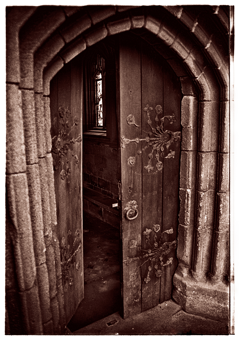 The ancient door of Tynemouth Priory — ornate ironwork scrolls on dark oak, a stained glass window glimpsed through the gap, stone archway framing centuries of passage
