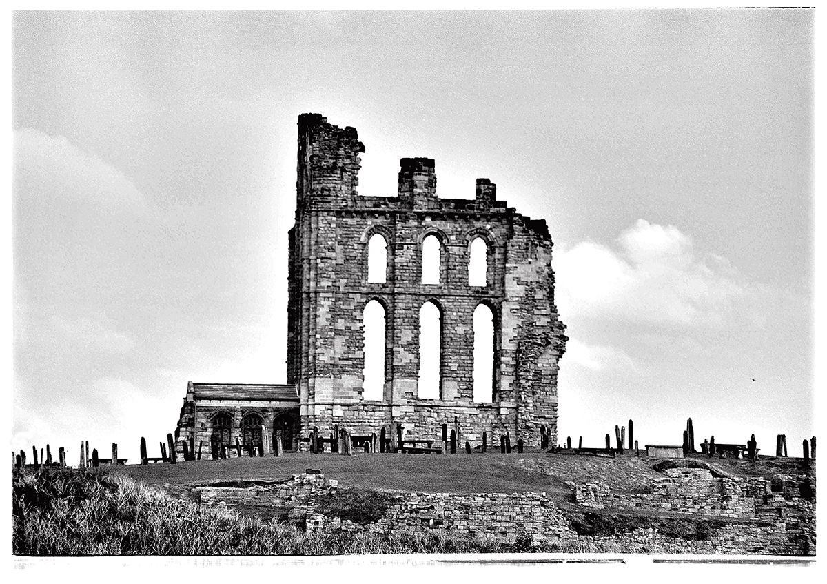 Tynemouth Priory ruins in black and white — the east window stands alone against the sky, gravestones in the foreground